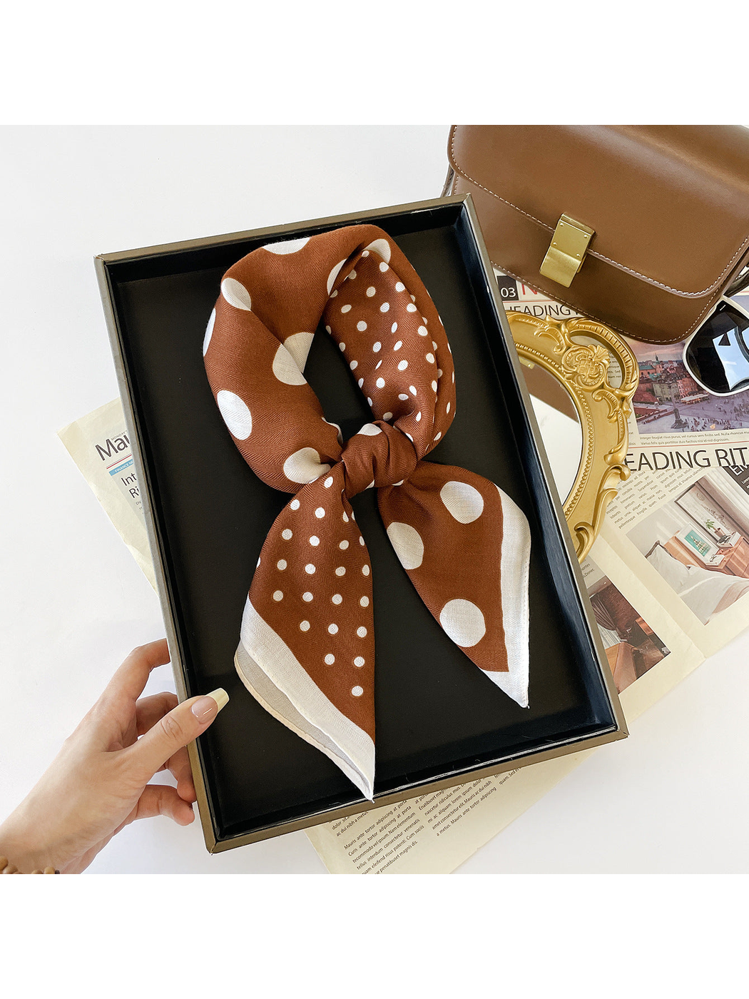 Brown scarf with white polka dots and a white border tied into a loop in a hand-held black tray.