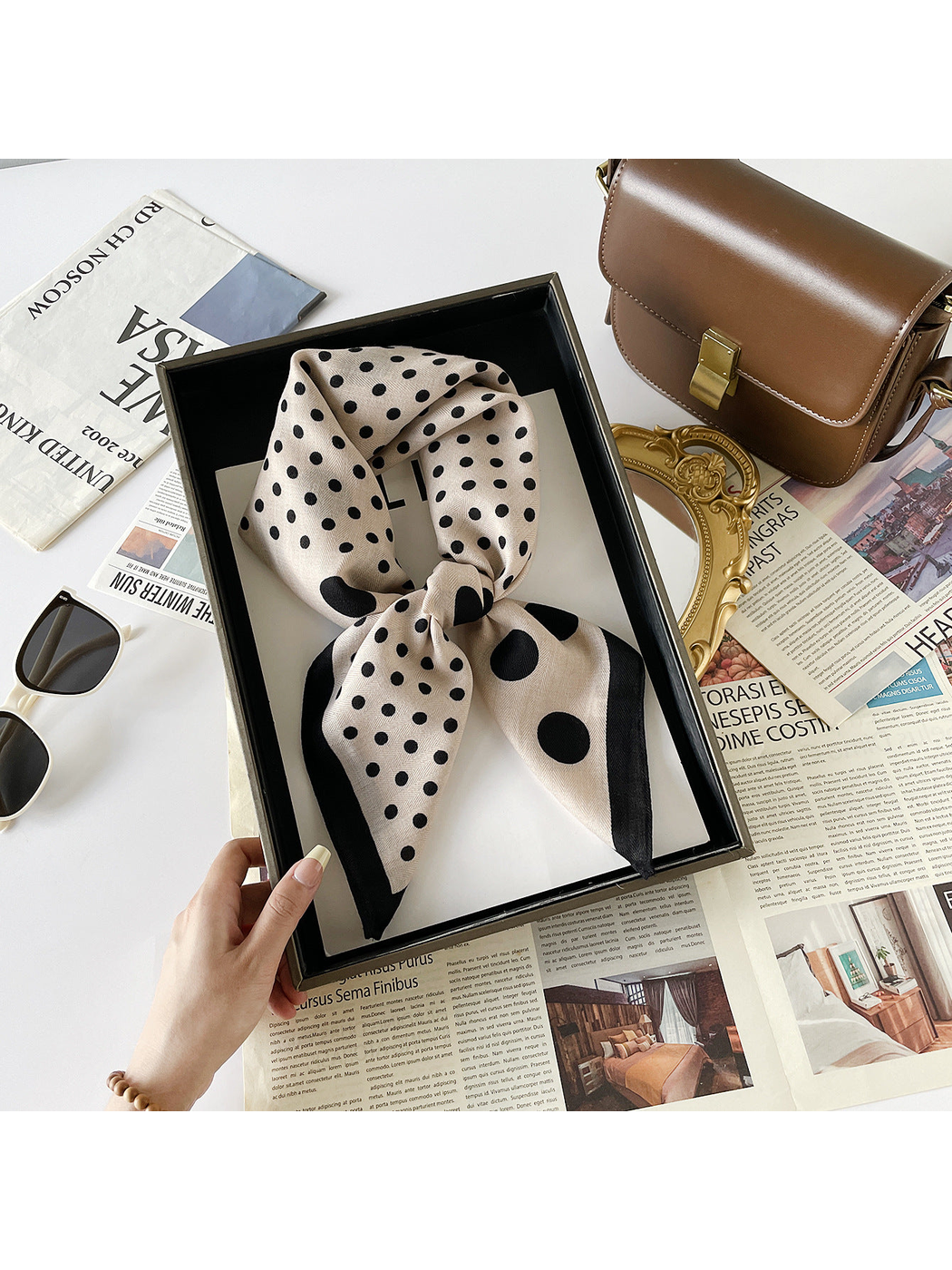Beige scarf with black polka dots and a black border tied into a loop in a hand-held tray.