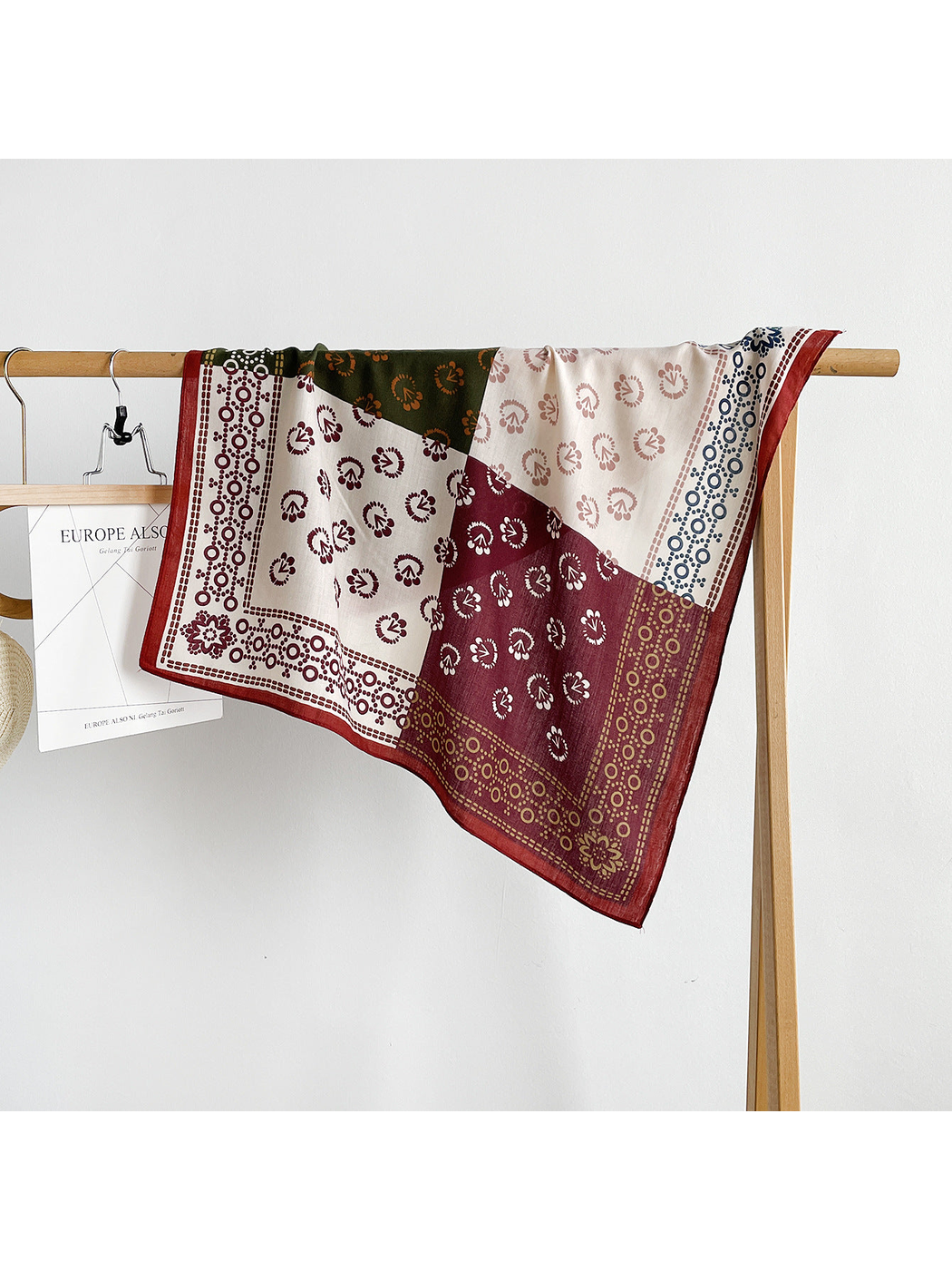 Burgundy square scarf draped over a rail, revealing contrasting panels and patterned borders.
