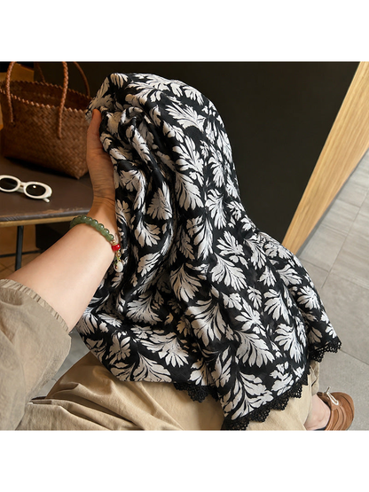 Multicolor botanical leaf-print scarf held and draped over a seated person's lap, with a lace-trimmed edge visible