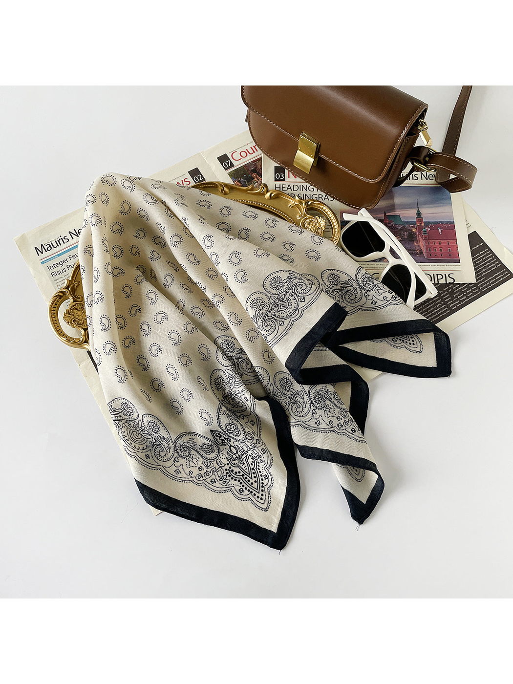 Black paisley square scarf draped beside a handbag, mirror, sunglasses, and magazine
